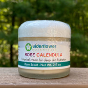Jar of Rose Calendula cream with Elderflower Botanicals label on a wooden surface with greenery in the background