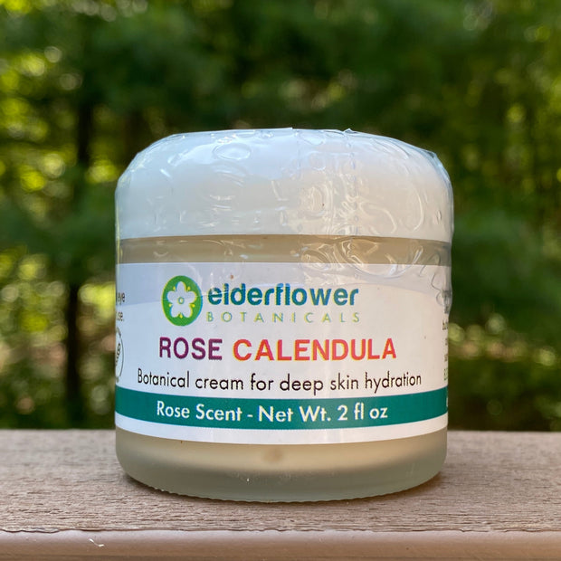 Jar of Rose Calendula cream with Elderflower Botanicals label on a wooden surface with greenery in the background